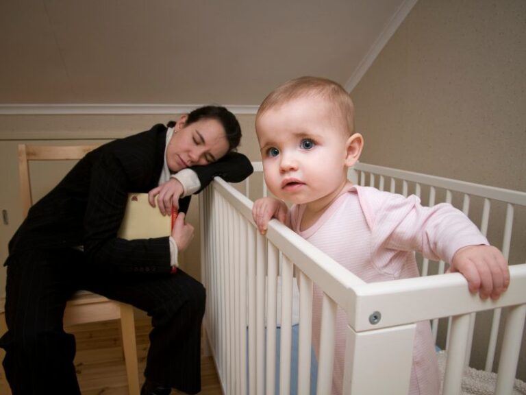 Mom is alseep leaning on the cot while her baby is wide awake with a different sleep pattern